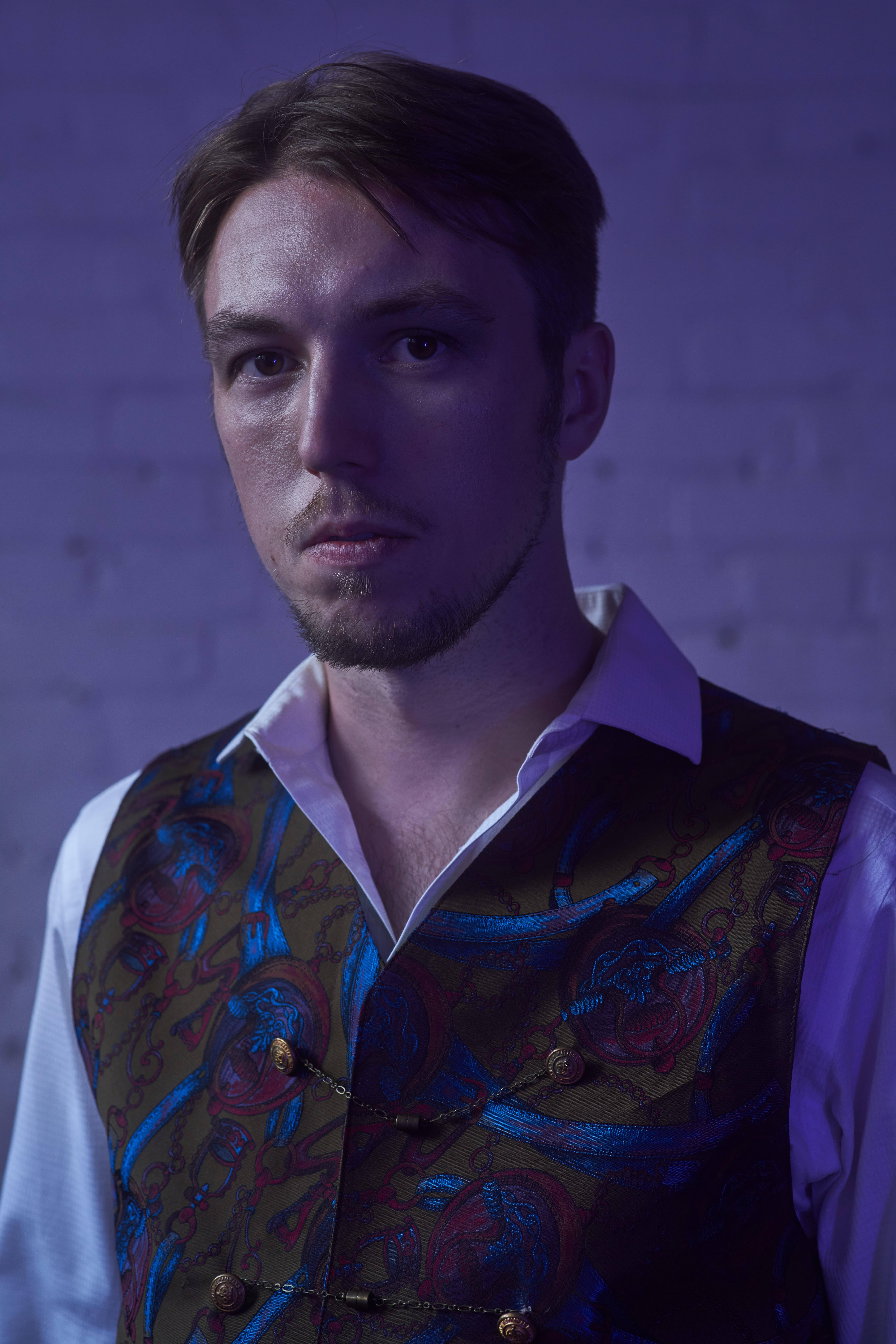 A colour photo of Tristan Zaba, standing in front of a white brick wall. The image has a purple tint to it.