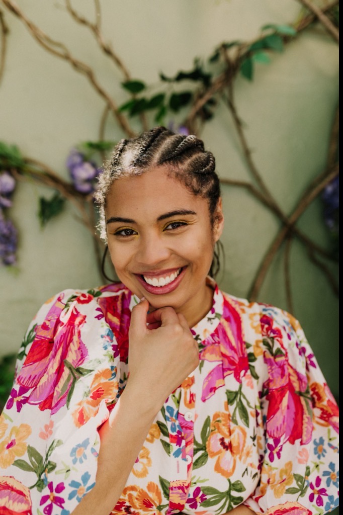 Picture is of a young woman with a big smile with teeth and large Fulani braids wearing a vibrant floral shirt in front of a wall covered in vines