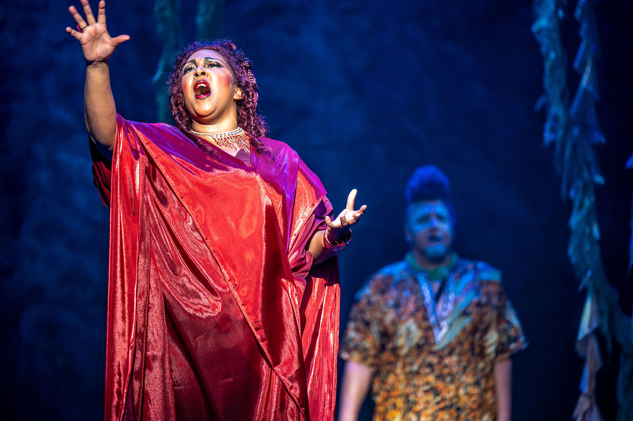 Of The Sea | Tapestry Opera Chantal Nurse as Serwa stands with her mouth open, mid-song. Her right arm is outstretched. She is wearing a shiny red gown.