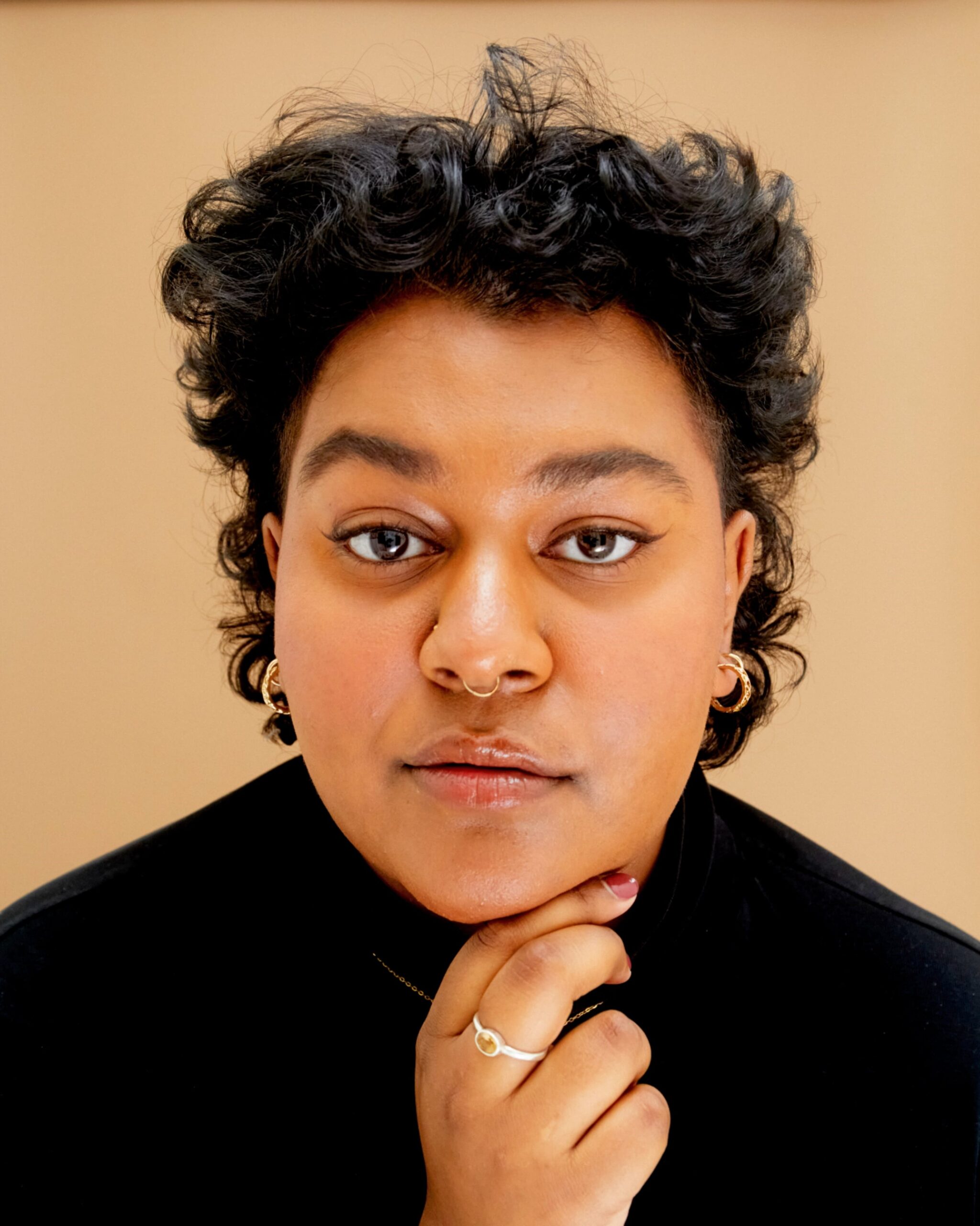 Anika is sitting close to the camera against a light brown background, facing forward with a slight smirk and their right hand on their chin. They have short, black, curly hair and are wearing a black turtle neck. They have one eyebrow slightly raised and are wearing various pieces of gold jewelry.