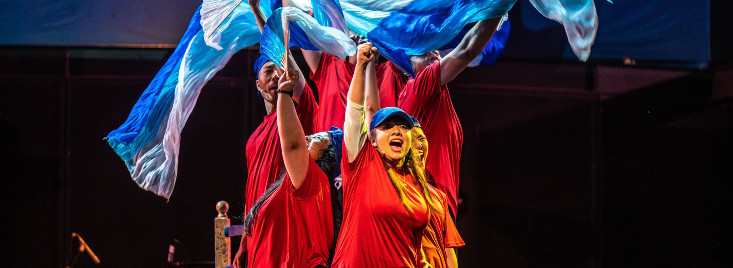 Dragon's Tale | Tapestry Opera Cast, wearing red shirts, wave blue and white flags in air