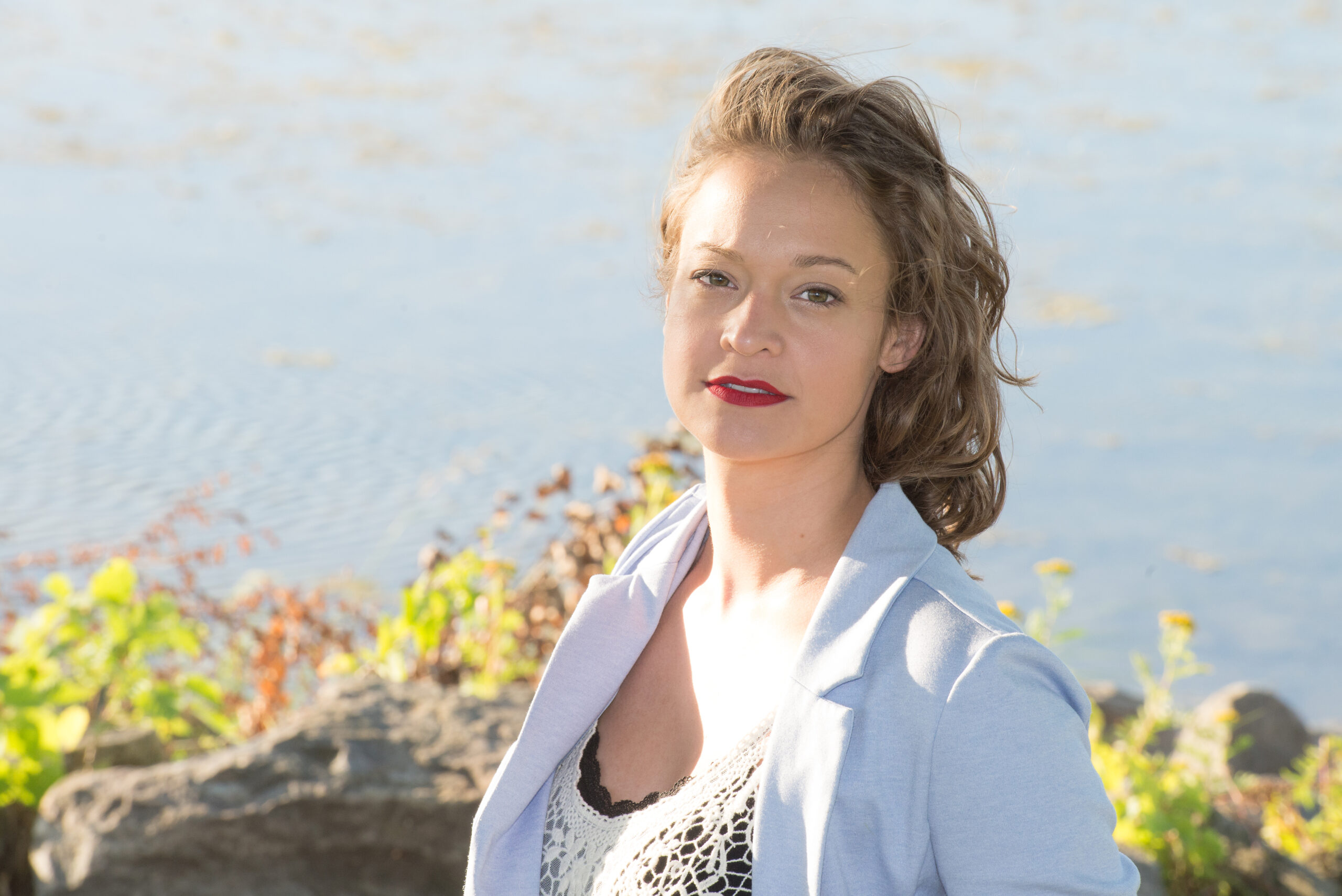 Composer Sophie Dupuis standing in front of a shoreline. There is water and some green foliage in the background. Sophie is has long blonde hair and is wearing a white suit jacket.