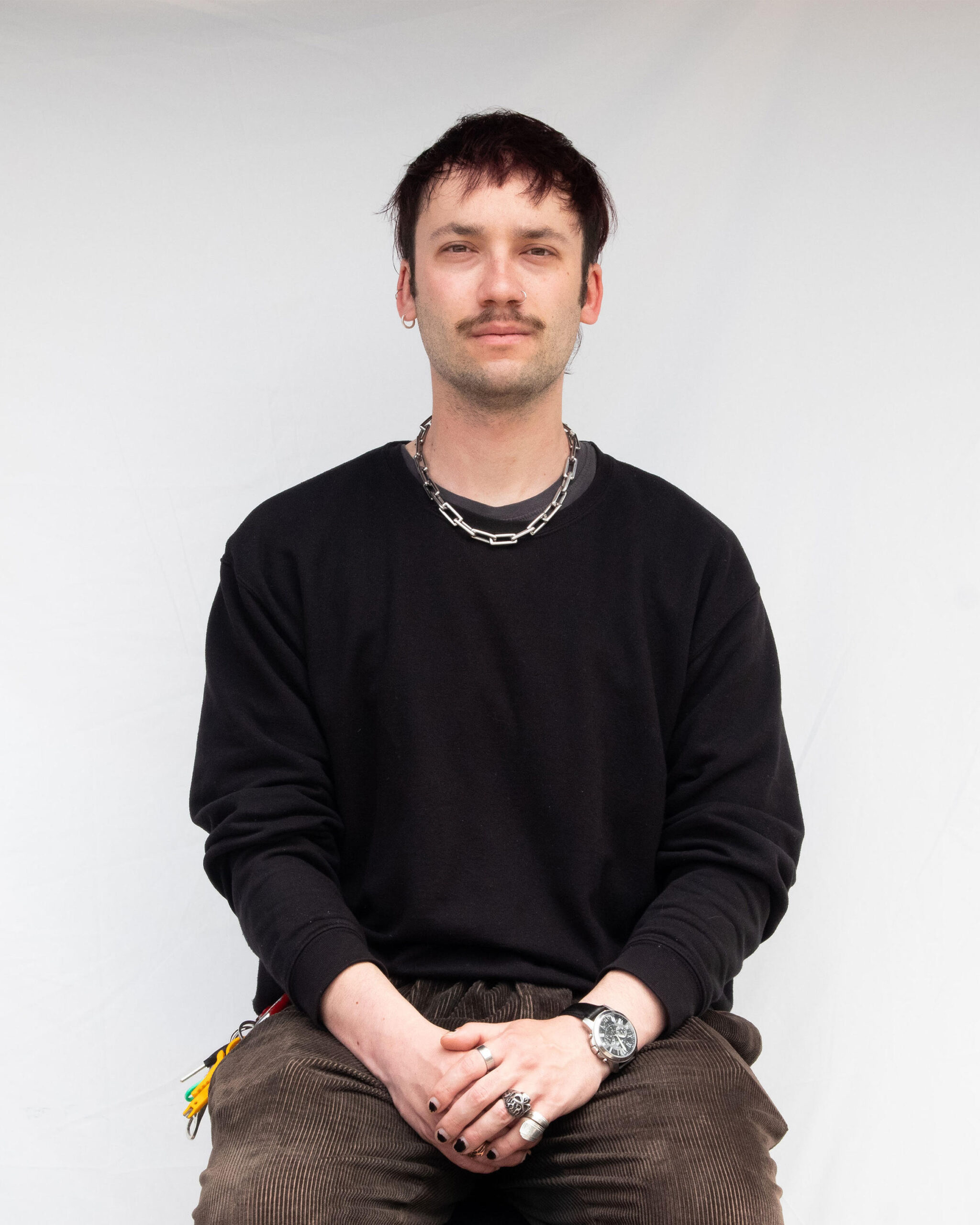 A picture of Nathan Bruce in a black sweater and dark brown corduroy pants. A chain hangs around his neck, and his hands are crossed over one another in his lap.