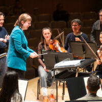 Conducting Fellow Juliane Gallant stands on the conducting podium, in profile. Maestro Gustavo Gimeno stands behind her to her left.